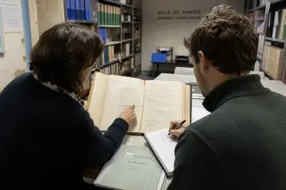 A municipal worker and a citizen examine council documents together in a subdued archive room, hands pointing at notes, with shelves of official binders in the background.