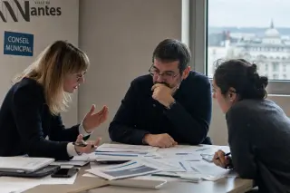 Three individuals deeply engaged in discussion over policy documents in a Nantes city hall meeting room, captured with soft lighting and a candid, documentary feel.