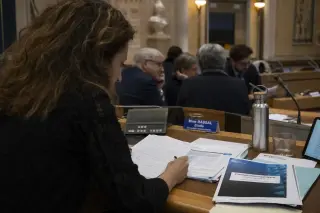 Someone reviewing notes and council papers in Nantes City Hall before a municipal meeting, with others focused in the background under gentle institutional lighting, atmosphere calm and engaged.