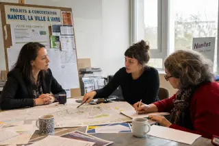 Three people in a Nantes municipal meeting room, focused on a collaborative discussion around city plans, in natural daylight, with an authentic, documentary feel.
