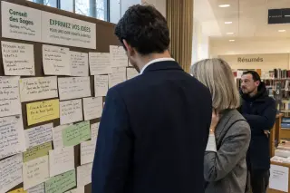 Three people interact near a community feedback board in a Nantes library, absorbed in reviewing handwritten notes, in a softly lit, candid setting.
