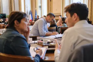Council chamber scene with a councillor focused on notes and two participants in a quiet discussion, natural light, authentic engaged atmosphere.