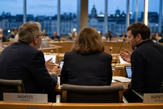 Three people seen from behind in a council chamber, deeply engaged in discussion, papers and tablets on the table, evening city lights visible through a glass wall.