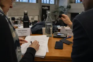 Council meeting desk with agenda, microphone, two participants partially visible, subdued institutional lighting, candid atmosphere.