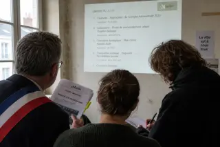 Three people in a Nantes municipal meeting room, intently reviewing a projected agenda together, natural daylight and focused, serious expressions highlighting a moment of civic engagement.