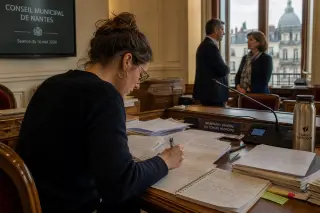 Municipal clerk in Nantes council chamber writing notes, with two officials talking quietly in the background near a window.