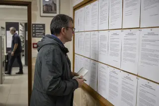 Citizen reading council documents on a bulletin board in a municipal archive room, absorbed in the details, with a municipal worker moving in the background.