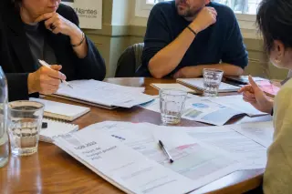 Three people in a city council room closely examining budget documents in serious discussion, with natural daylight and an intimate focus at table level.
